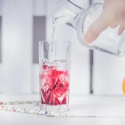 Ręka wlewa wodę z dzbanka do szklanki z lodem i czerwonym napojem. Obok pomarańcza.