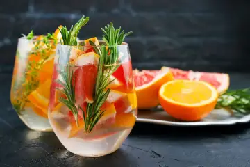 Orzeźwiający drink z grejpfrutem, pomarańczą i rozmarynem. Dwa szklane kieliszki wypełnione wodą, owocami i ziołami.